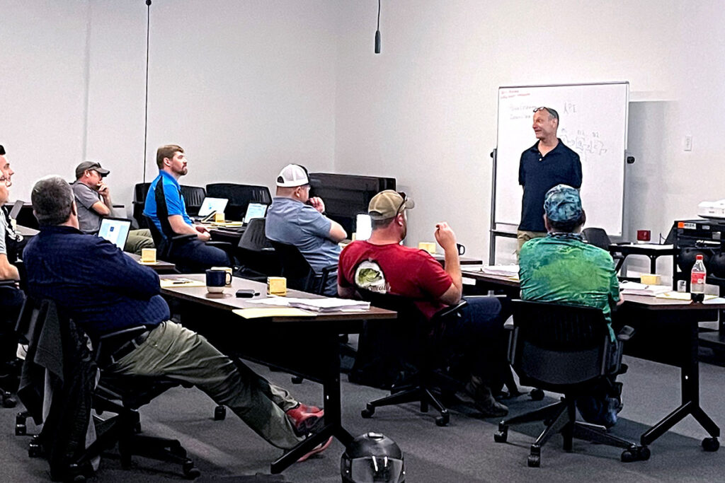 Several adult male students in classroom with teacher standing in front of white board.